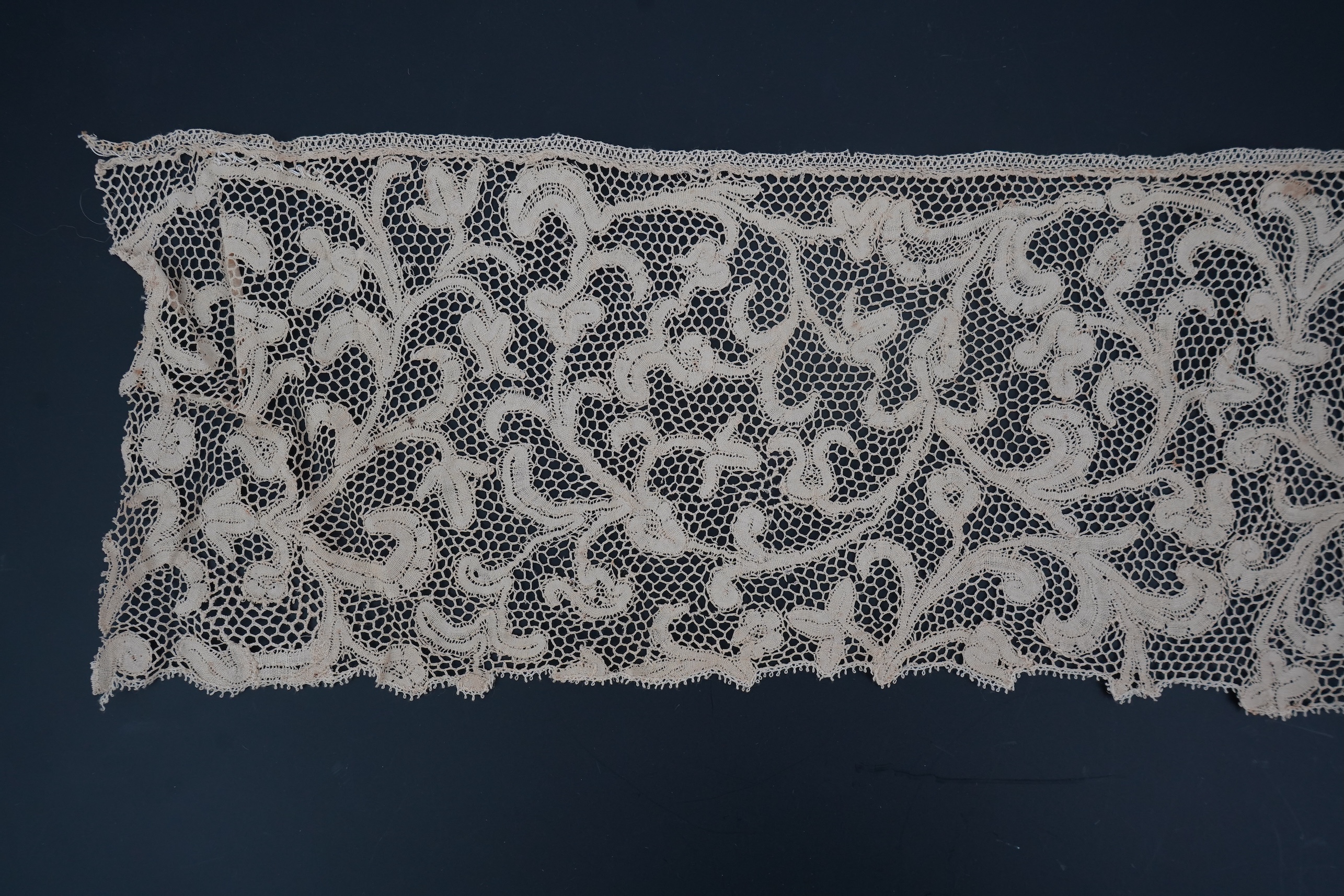 A length of 1740-1760 Italian, Milanese bobbin lace flounce, made from flat braiding or tapes, placed into scrolling patterns, attached to brides, made into a honeycomb mesh. 252cm long x 16cm deep. Condition - one small